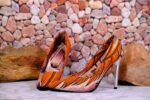 Handmade African wax print high-heeled pumps with silver stiletto, orange and black pattern, on marble surface with river stones.