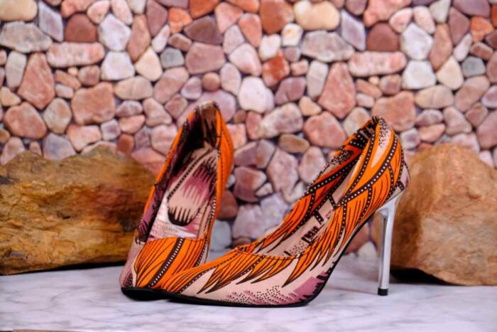 Handmade African wax print high-heeled pumps with silver stiletto, orange and black pattern, on marble surface with river stones.