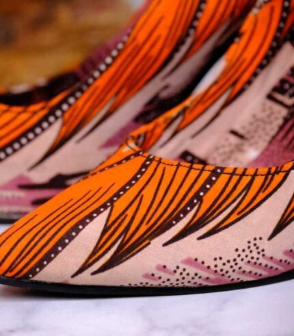 Handmade African wax print high-heeled pumps with silver stiletto, orange and black pattern, on marble surface with river stones.