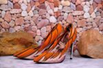Handmade African wax print high-heeled pumps with silver stiletto, orange and black pattern, on marble surface with river stones.