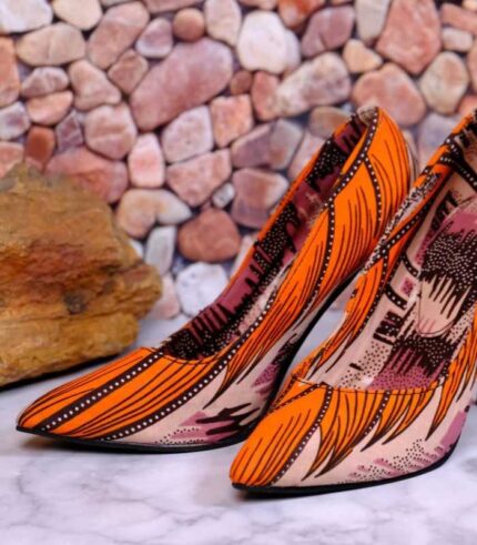 Handmade African wax print high-heeled pumps with silver stiletto, orange and black pattern, on marble surface with river stones.