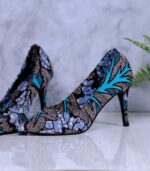 Handmade African wax print high-heeled pumps in blue, turquoise, and white with black accents, displayed on a reflective surface with a drum and plant.