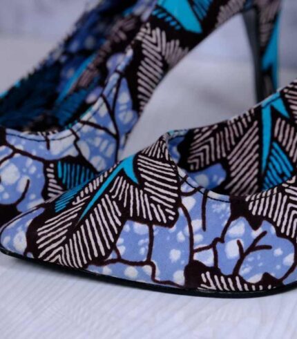 Handmade African wax print high-heeled pumps in blue, turquoise, and white with black accents, displayed on a reflective surface with a drum and plant.