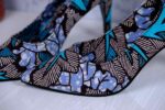 Handmade African wax print high-heeled pumps in blue, turquoise, and white with black accents, displayed on a reflective surface with a drum and plant.
