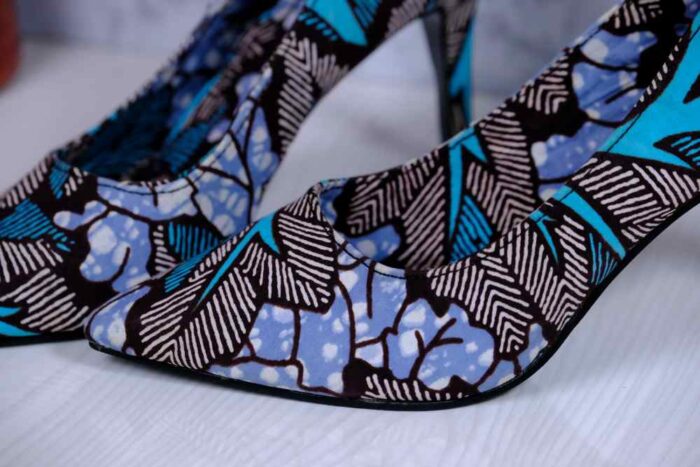 Handmade African wax print high-heeled pumps in blue, turquoise, and white with black accents, displayed on a reflective surface with a drum and plant.
