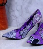 Handmade African print high-heeled shoes in purple with black, white, and pink patterns, displayed against a marble wall with plant leaves.