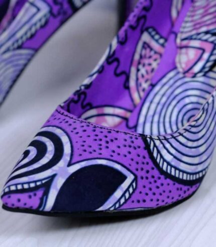 Handmade African print high-heeled shoes in purple with black, white, and pink patterns, displayed against a marble wall with plant leaves.