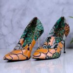 Handmade African print high-heeled shoes in orange, green, and black pattern with a wooden drum and plant in the background.