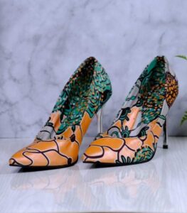 Handmade African print high-heeled shoes in orange, green, and black pattern with a wooden drum and plant in the background.