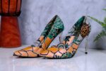Handmade African print high-heeled shoes in orange, green, and black pattern with a wooden drum and plant in the background.