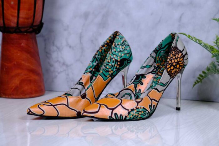 Handmade African print high-heeled shoes in orange, green, and black pattern with a wooden drum and plant in the background.