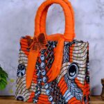 Adire handbag with bold orange, black, and white print, twisted orange handle, and shimmering bow detail. A stylish, handcrafted African accessory.