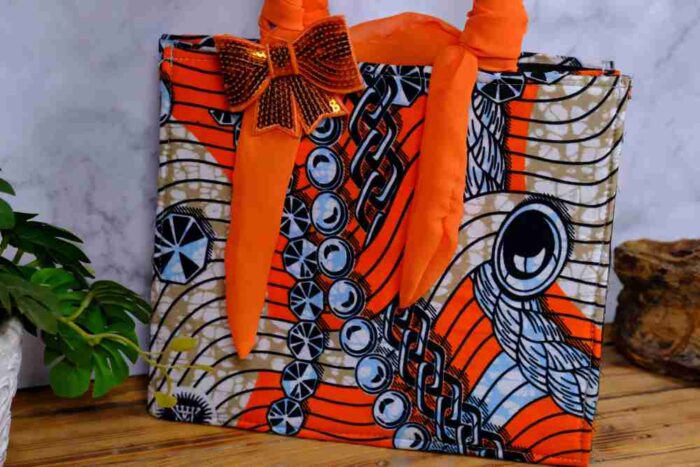 Adire handbag with bold orange, black, and white print, twisted orange handle, and shimmering bow detail. A stylish, handcrafted African accessory.