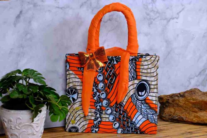 Adire handbag with bold orange, black, and white print, twisted orange handle, and shimmering bow detail. A stylish, handcrafted African accessory.