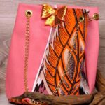 Desert Mirage Cinch Bag featuring pink faux leather, orange Ankara feather motif, gold-sequin bow, and gold chain strap on a wooden background.