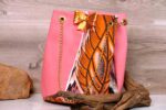 Desert Mirage Cinch Bag featuring pink faux leather, orange Ankara feather motif, gold-sequin bow, and gold chain strap on a wooden background.