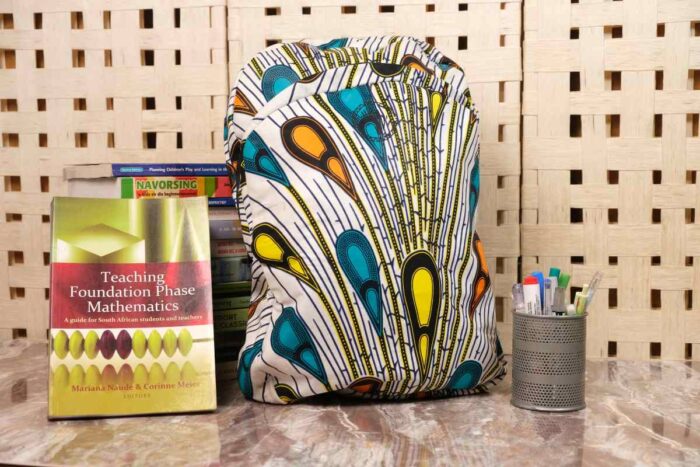 Plume Ankara Backpack with feather-inspired teardrop motifs in teal, golden yellow, and amber orange on ivory base, featuring L’Africaine Boutique logo.