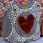Red Heart Statement Bag with black and white patterned fabric, red sequined heart, and gold chain handles. Displayed against a stone wall with rocks.
