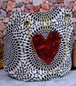Red Heart Statement Bag with black and white patterned fabric, red sequined heart, and gold chain handles. Displayed against a stone wall with rocks.