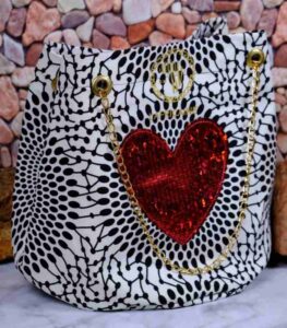 Red Heart Statement Bag with black and white patterned fabric, red sequined heart, and gold chain handles. Displayed against a stone wall with rocks.