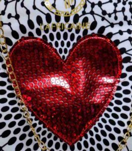 Red Heart Statement Bag with black and white patterned fabric, red sequined heart, and gold chain handles. Displayed against a stone wall with rocks.