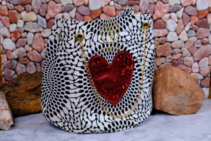 Red Heart Statement Bag with black and white patterned fabric, red sequined heart, and gold chain handles. Displayed against a stone wall with rocks.