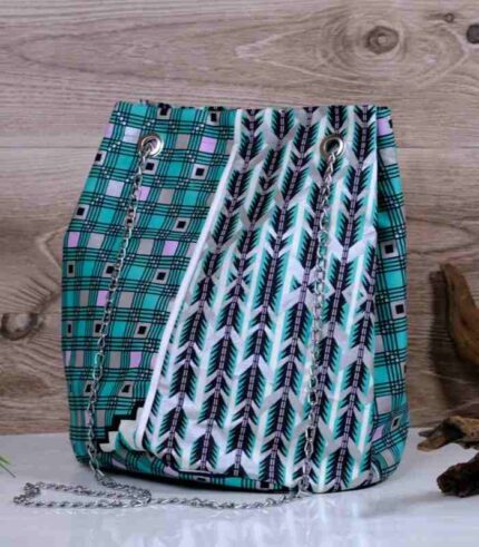 Teal Geometry Cinch Bag with teal, black, and white geometric print, silver chain strap, and drawstring closure. Displayed with plant and driftwood decor.