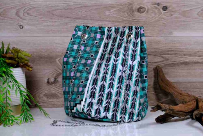 Teal Geometry Cinch Bag with teal, black, and white geometric print, silver chain strap, and drawstring closure. Displayed with plant and driftwood decor.