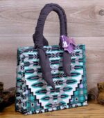 Teal Geometry Handbag with geometric teal, black, and white pattern, dark fabric handles, and a purple bow. Displayed on wooden planks with driftwood.