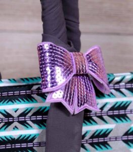 Teal Geometry Handbag with geometric teal, black, and white pattern, dark fabric handles, and a purple bow. Displayed on wooden planks with driftwood.