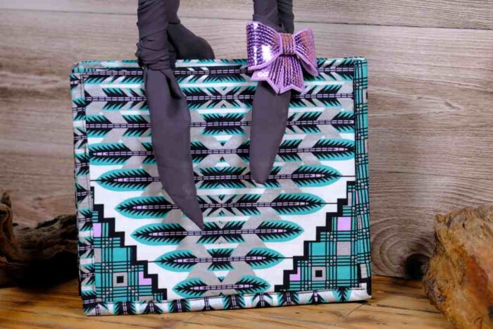 Teal Geometry Handbag with geometric teal, black, and white pattern, dark fabric handles, and a purple bow. Displayed on wooden planks with driftwood.