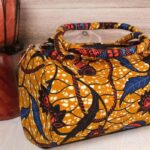 Mustard Ankara handbag in wax fabric with navy, red, and black floral and leaf patterns, padded handles, and gold zip closure.