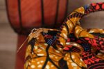 Mustard Ankara handbag in wax fabric with navy, red, and black floral and leaf patterns, padded handles, and gold zip closure.
