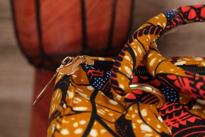 Mustard Ankara handbag in wax fabric with navy, red, and black floral and leaf patterns, padded handles, and gold zip closure.