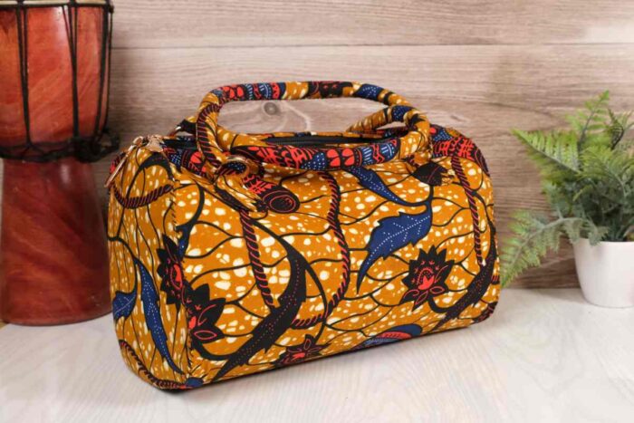 Mustard Ankara handbag in wax fabric with navy, red, and black floral and leaf patterns, padded handles, and gold zip closure.
