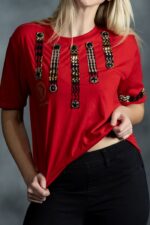 Red tee with vertical black and gold patterned strips and circular embellishments on chest and sleeves from L’Africaine Boutique.