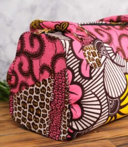 ankara floral swirl handbag in vibrant pink wax fabric with yellow, white, and brown floral swirl patterns, padded handles, gold rings, and top zipper.