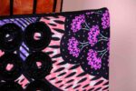 ankara midnight bloom clutch in purple, pink, and black Ankara wax fabric with hand-embroidered sequin rings, gold logo, and pink tassel zipper accent.