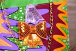 ankara sequin bucket bag in multicolor Ankara wax fabric with zigzag print, green faux leather trim, gold chain strap, and sequin bow embellishment.