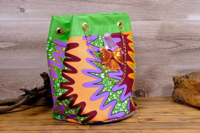 ankara sequin bucket bag in multicolor Ankara wax fabric with zigzag print, green faux leather trim, gold chain strap, and sequin bow embellishment.