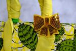 ankara sequin bow handbag in yellow and green Ankara wax fabric with leaf and pod motifs, padded handles, gold-tone rings, and gold sequin bow.