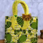 ankara sequin bow handbag in yellow and green Ankara wax fabric with leaf and pod motifs, padded handles, gold-tone rings, and gold sequin bow.