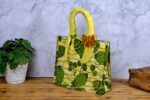 ankara sequin bow handbag in yellow and green Ankara wax fabric with leaf and pod motifs, padded handles, gold-tone rings, and gold sequin bow.