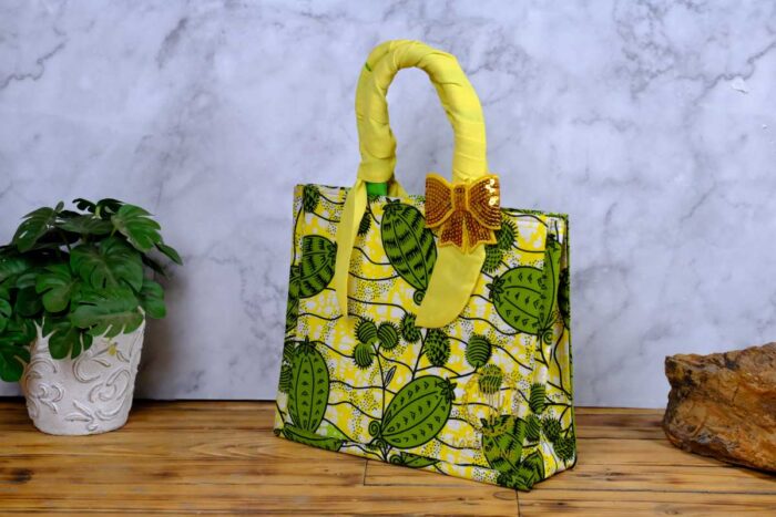 ankara sequin bow handbag in yellow and green Ankara wax fabric with leaf and pod motifs, padded handles, gold-tone rings, and gold sequin bow.