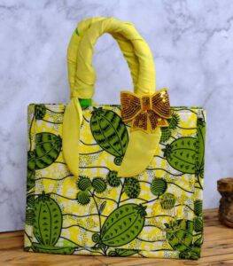 ankara sequin bow handbag in yellow and green Ankara wax fabric with leaf and pod motifs, padded handles, gold-tone rings, and gold sequin bow.