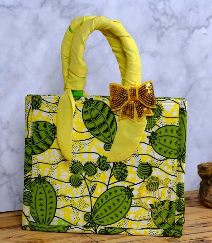 ankara sequin bow handbag ankara sequin bow handbag in yellow and green Ankara wax fabric with leaf and pod motifs, padded handles, gold-tone rings, and gold sequin bow.